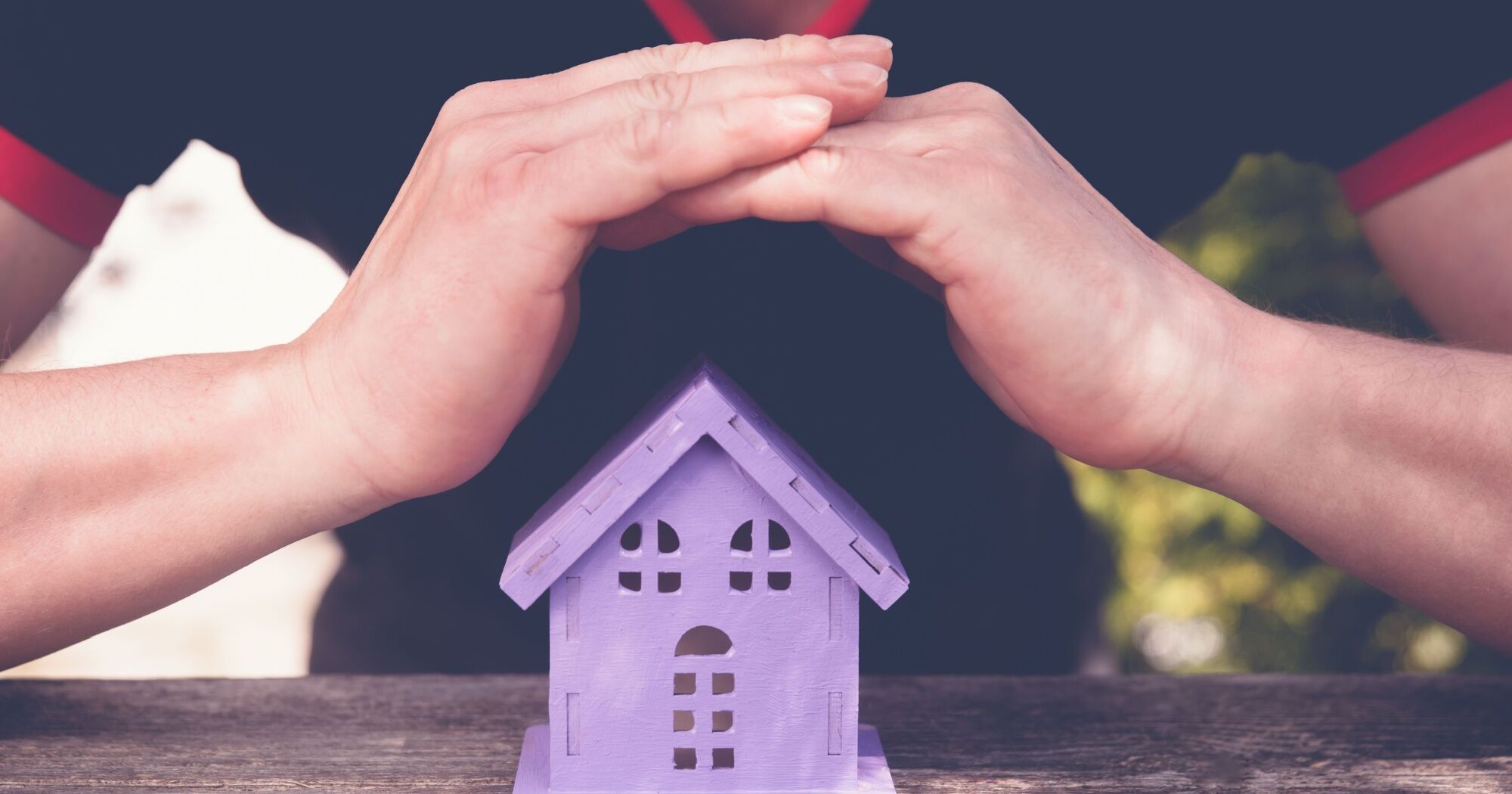 Hands protectively covering a small purple model house on a wooden surface, symbolizing the importance of roofing warranties in safeguarding homes.